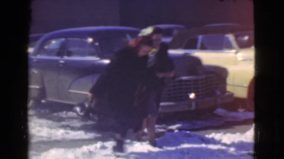 A woman in a dark coat bends toward the snowy ground near a dark sedan and a pale yellow car in Cleveland winter footage.