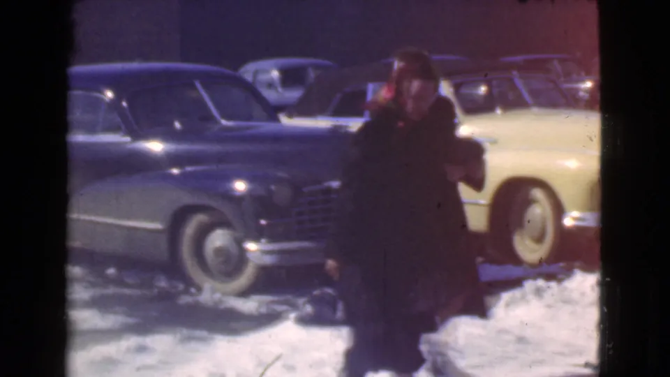 A woman moves across a snow-covered Cleveland parking area, with a dark sedan and pale yellow car behind her.