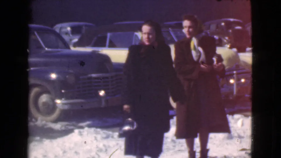 Two women in winter coats and hats face the camera in a snowy Cleveland parking lot, with late-1930s cars behind them.