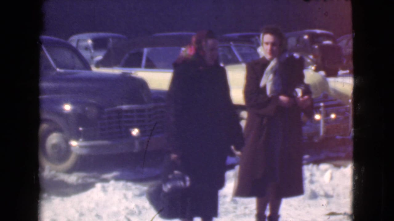 Two women in long winter coats and headscarves appear near parked prewar cars on a snowy Cleveland street, consistent with the 1939 home-movie metadata.