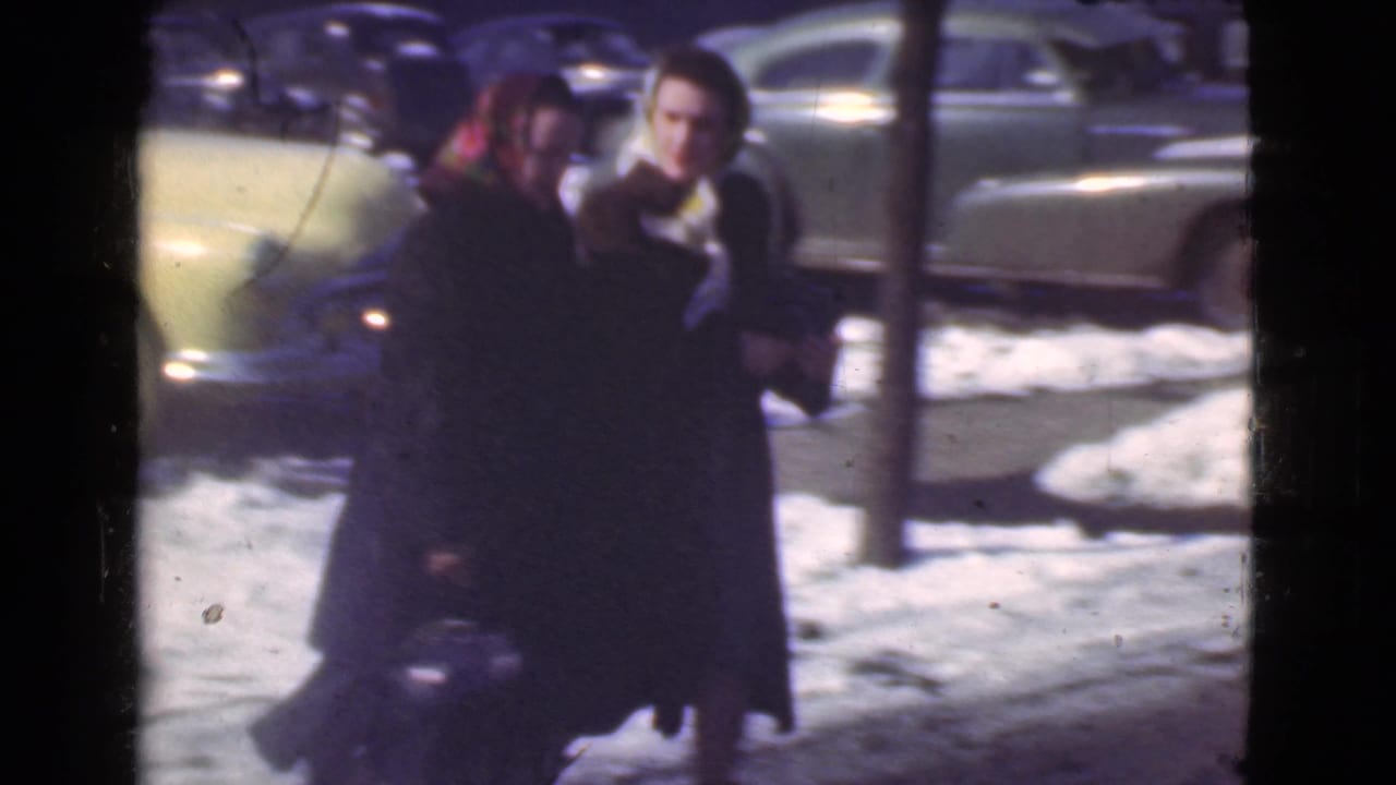 Two women in dark coats walk together across snow with parked cars behind them.