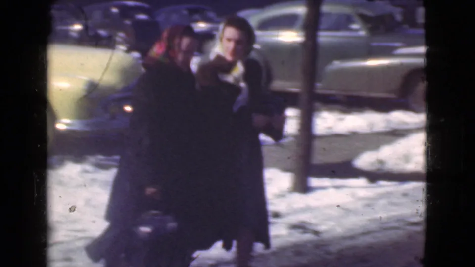 Two women in dark winter coats move across a snow-covered street in Cleveland, with late-1930s automobiles parked behind them.