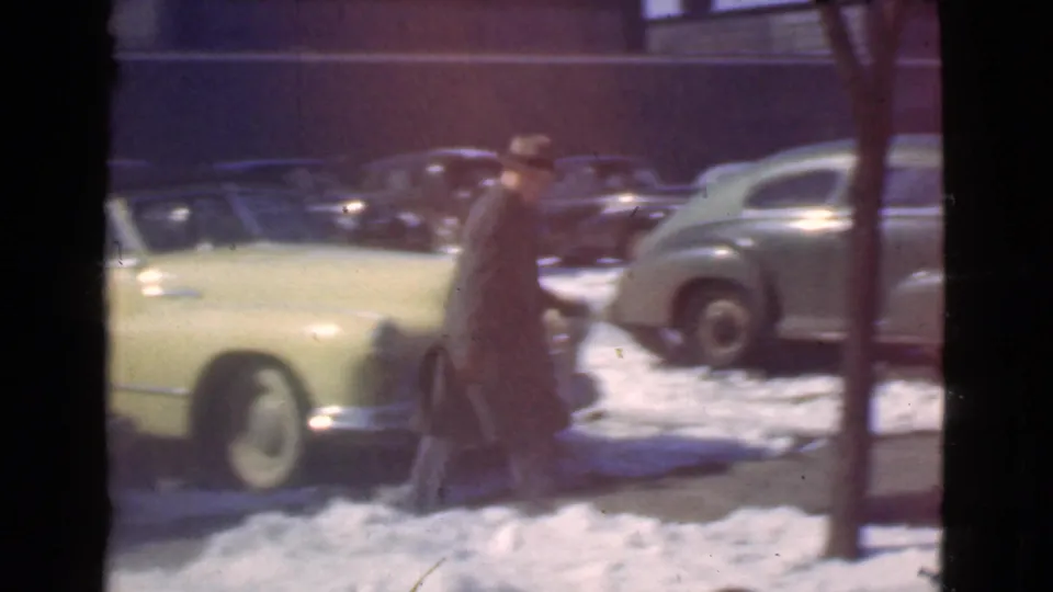 A man in a brimmed hat and long coat walks past a pale yellow automobile on a snowy Cleveland street, with more parked cars in the background.
