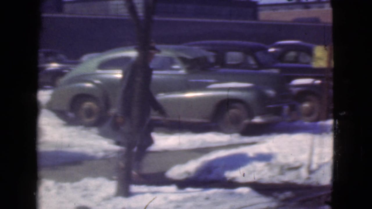 Snowy street with parked vintage cars and a blurred pedestrian at the left edge.