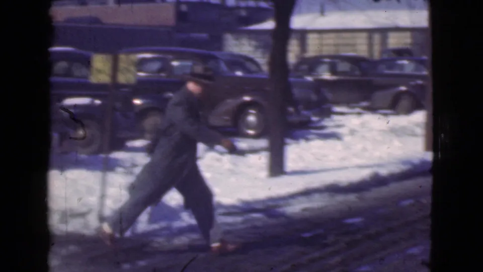 A man in a dark overcoat and fedora walks briskly across a snowy Cleveland street in 1939 with parked automobiles behind him.