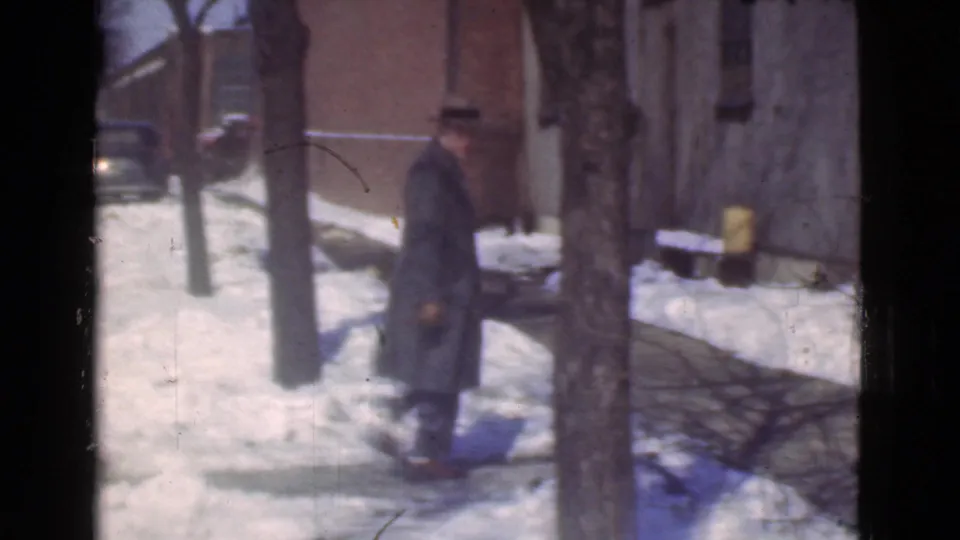 A man in a dark coat and hat appears near a tree and building wall on a snowy Cleveland sidewalk at the end of the short 1939 shot.
