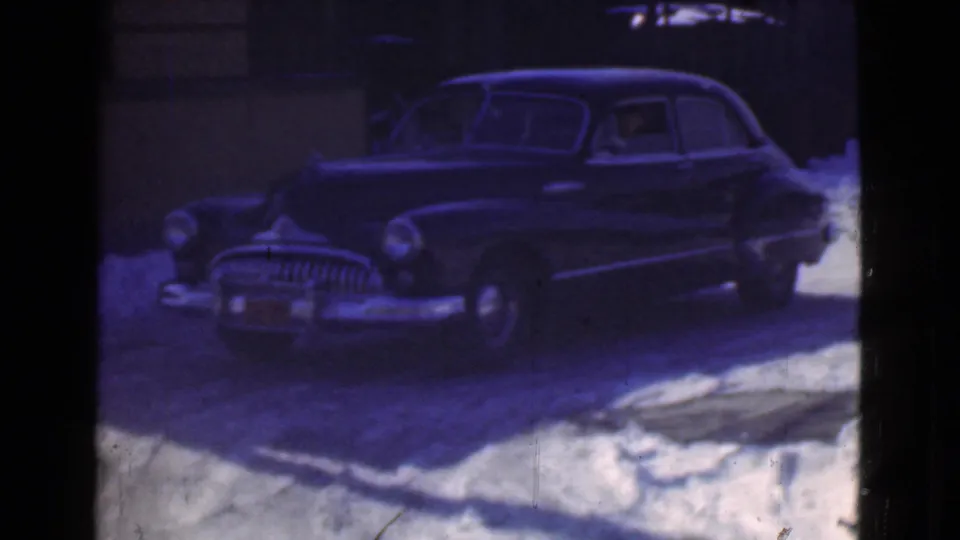 A dark rounded sedan sits near snowbanks on a Cleveland winter street, seen through soft, color-shifted 8mm film.