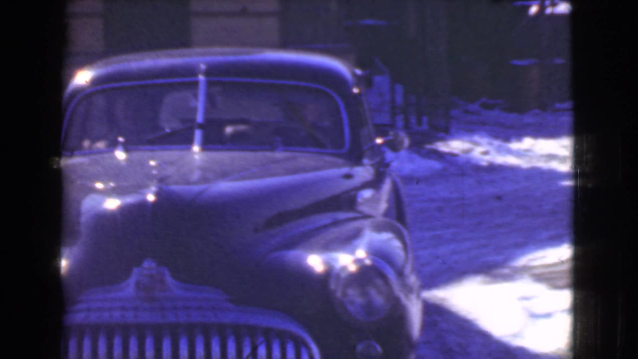 Vintage sedan approaching on a snow-dusted street with dark film borders.