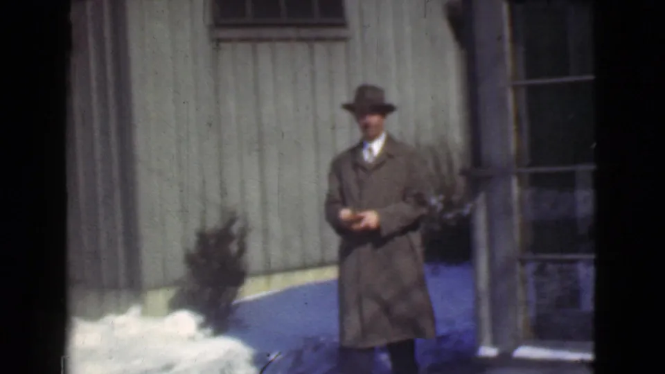 1939 Cleveland frame showing a man in a long coat and fedora outside a wooden building beside snow patches.