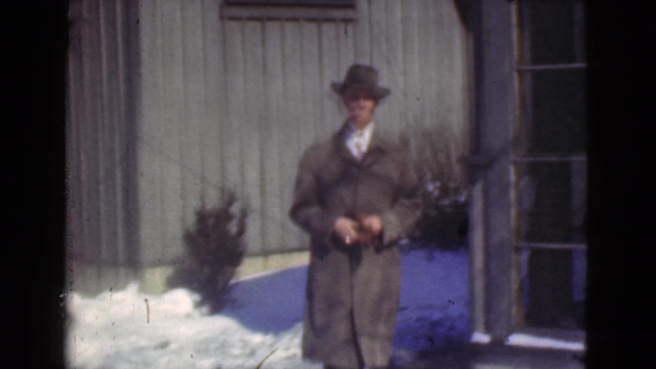 Man in a suit and fedora stands outside a wooden building with snow on the ground.