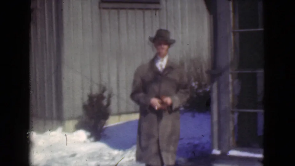 In 1939 Cleveland, a suited man in a fedora stands outside a wooden building beside patches of snow.