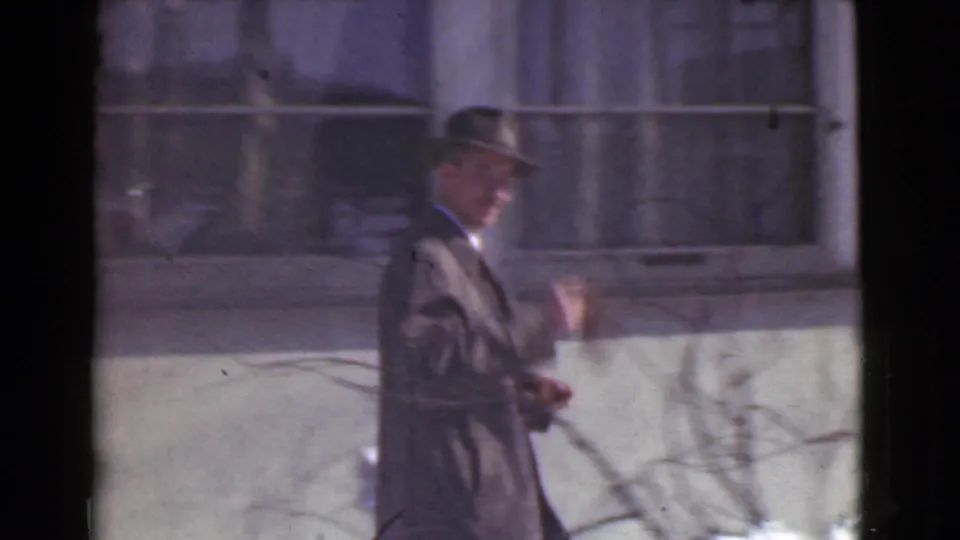 The suited man raises a hand in a friendly wave while walking by building windows in Cleveland, 1939.