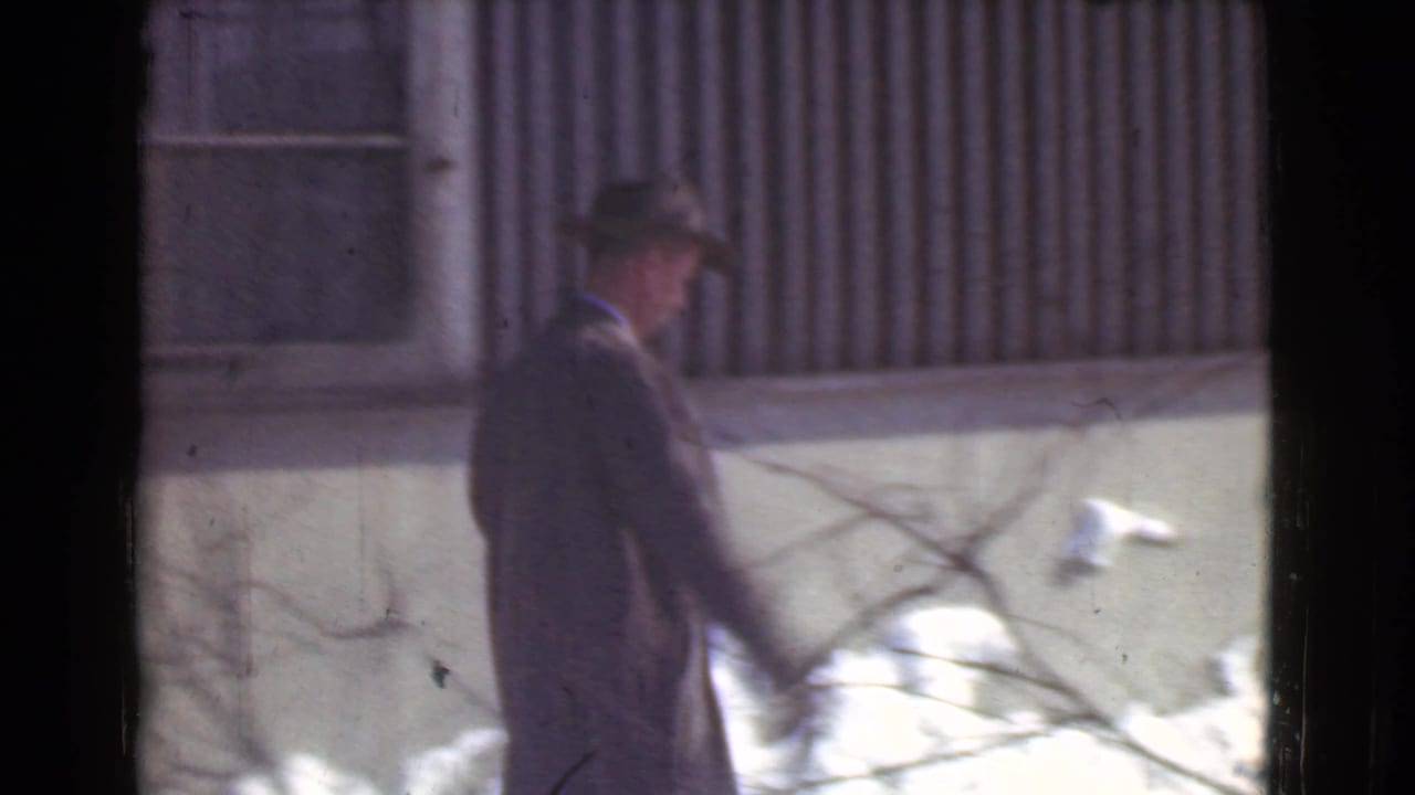 Man in a dark coat and fedora walks beside a snowy corrugated building.