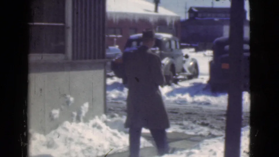 A man in a long dark coat walks away toward vintage automobiles on a snowy Cleveland street in the supplied 1939 setting.