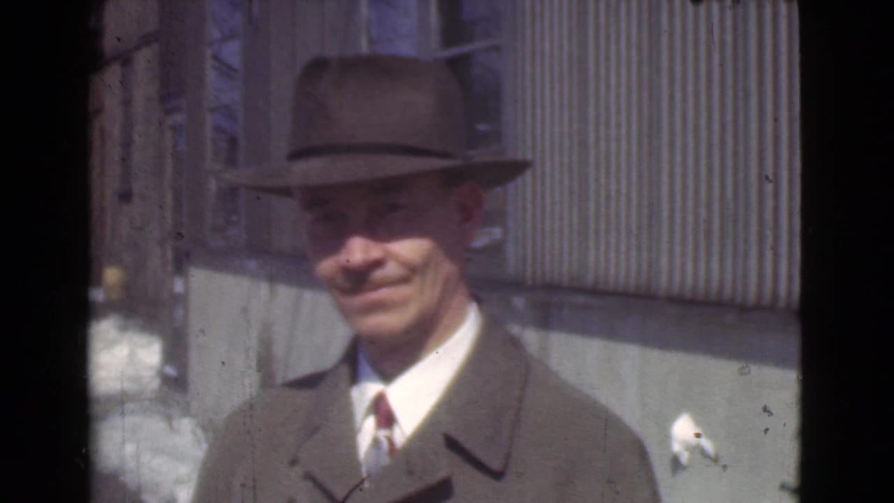 Adult man in a fedora and overcoat smiles outdoors beside a corrugated wall.