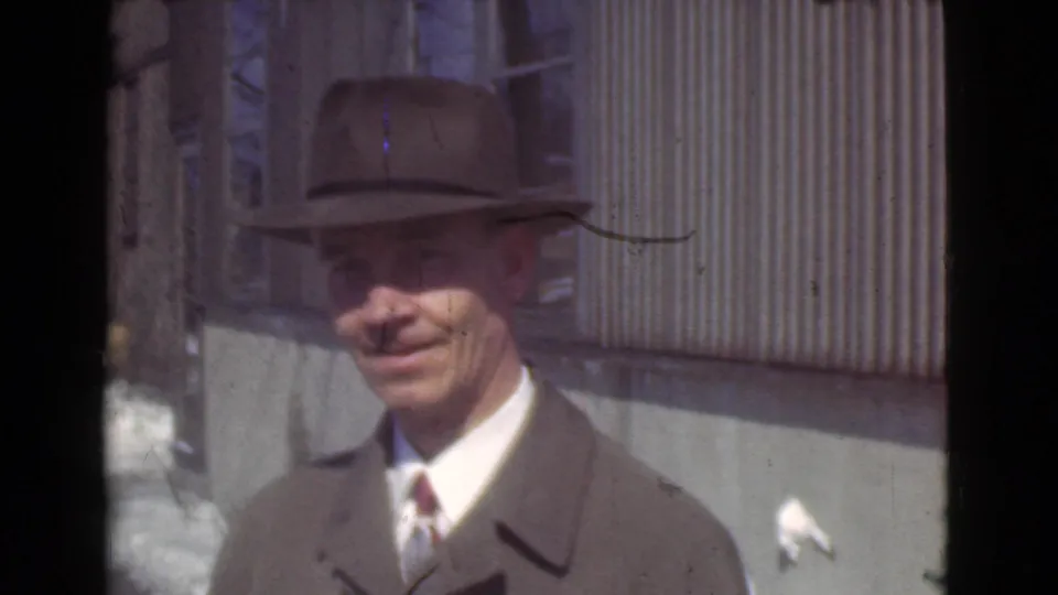 The man remains framed close to camera, smiling under the brim of his hat against a sunlit corrugated wall.