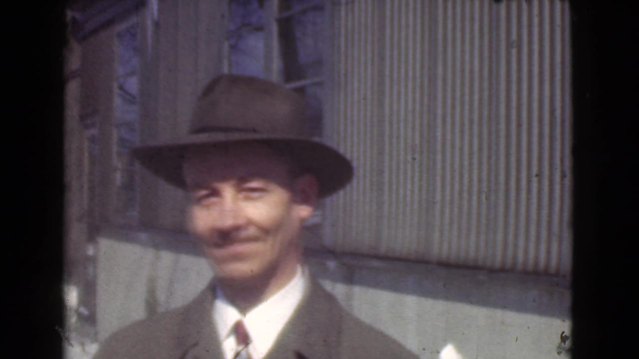 Adult man in a fedora and suit smiles near a corrugated wall.