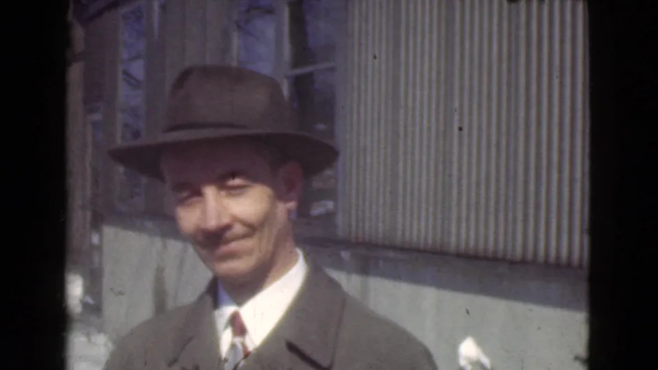 In the Cleveland 1939 home-movie scene, a man in a fedora and overcoat faces the camera with a slight smile.