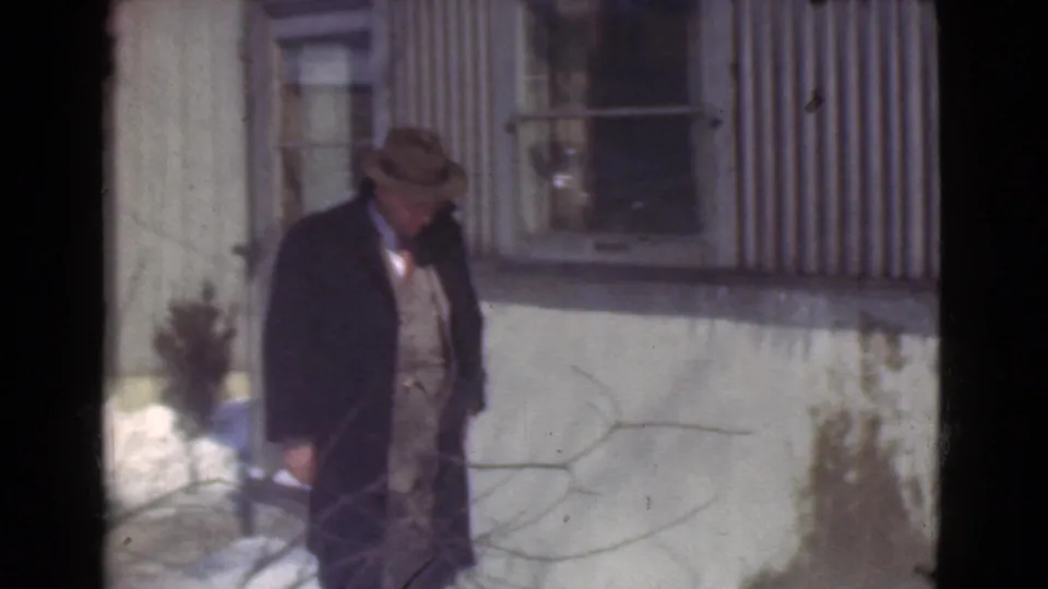 The man appears farther from the camera near the corrugated building, wearing a dark overcoat over a suit and vest.
