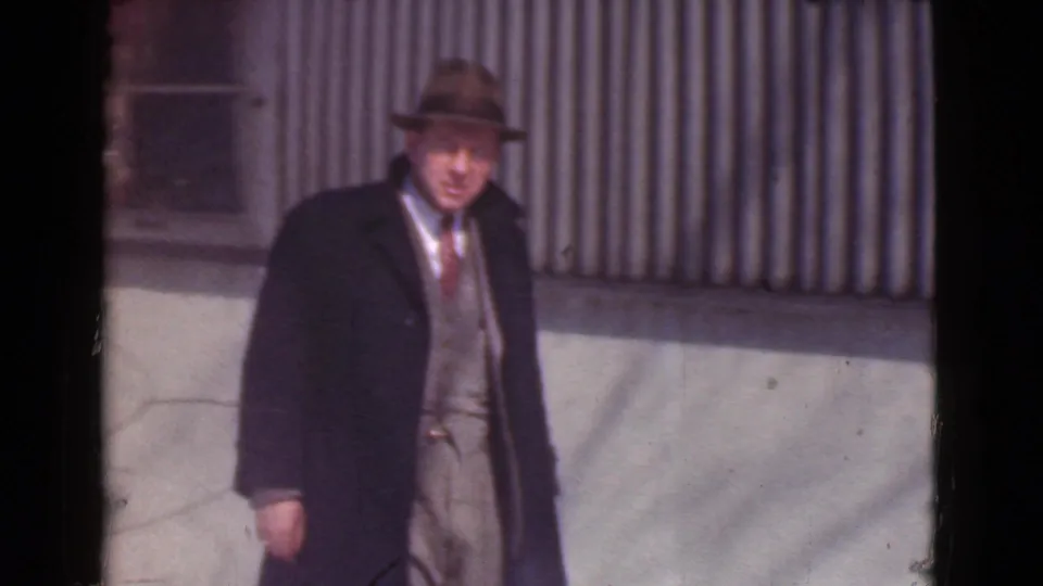 The 1939 Cleveland home-movie frame shows the man walking near the building in a fedora, overcoat, vest, and tie.