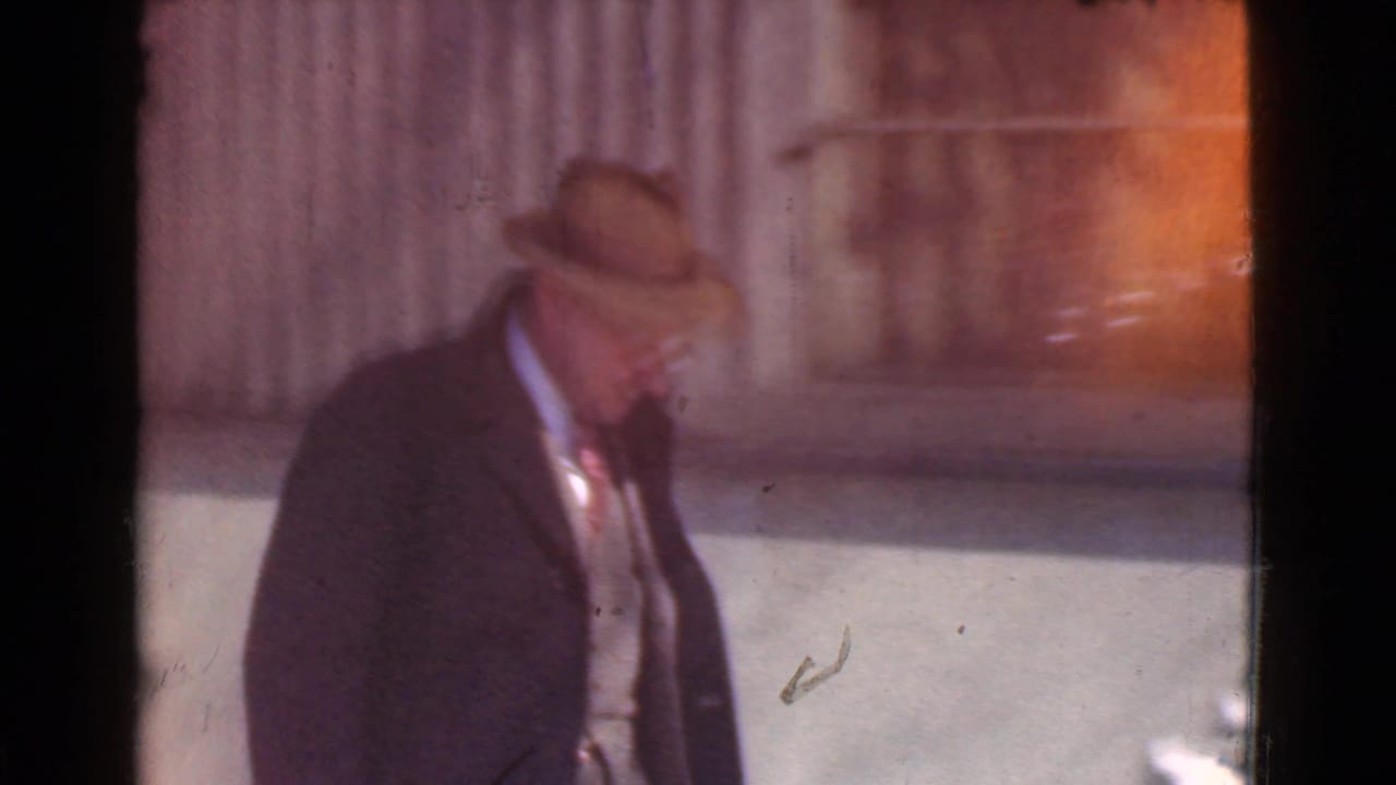 Man in a dark coat and fedora walking beside a snowy street.