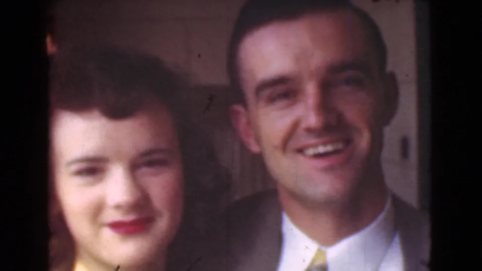 A smiling adult couple appears in a close home-movie portrait, with faces softly focused and warm indoor or doorway light.