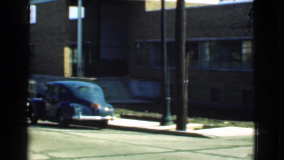 The 1939 Cleveland street scene shows a dark rounded car parked near a brick building, with windows, poles, sidewalk, and pavement visible.