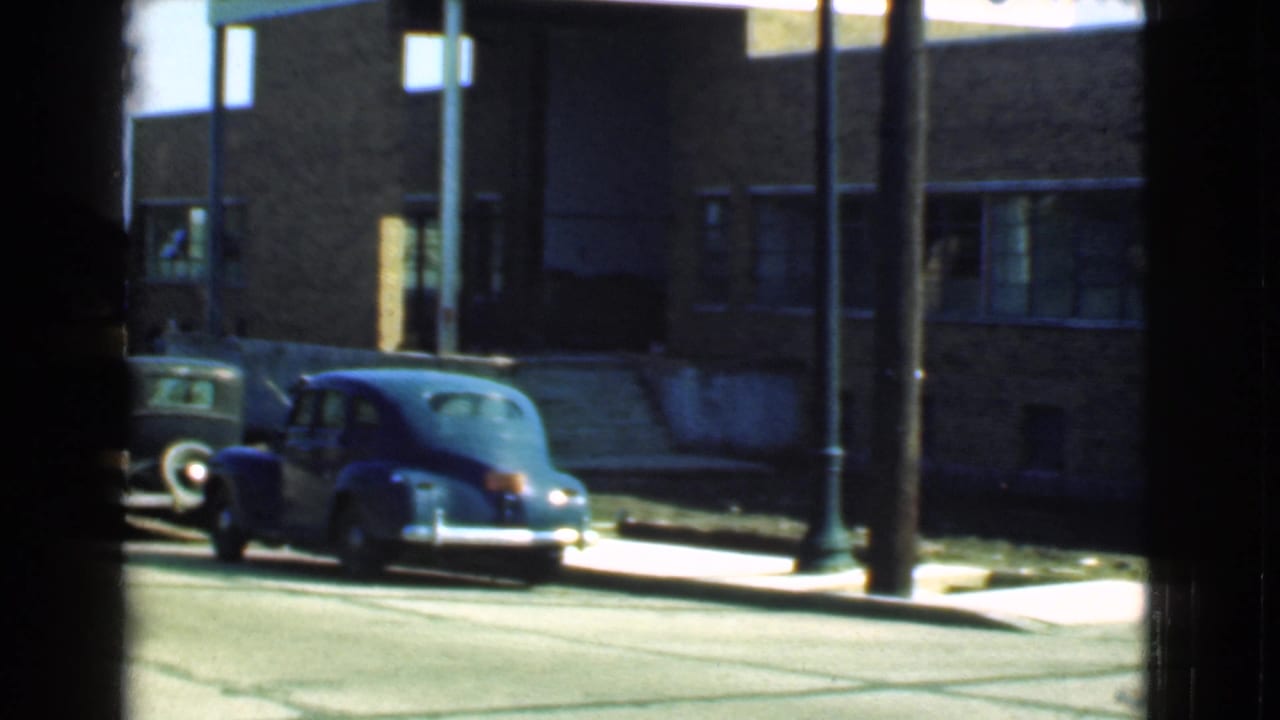 1939 Cleveland street view with a rounded blue sedan parked near a brick building and street posts, partly framed by a window.