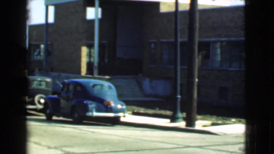 1939 Cleveland street view with a rounded blue sedan parked near a brick building and street posts, partly framed by a window.
