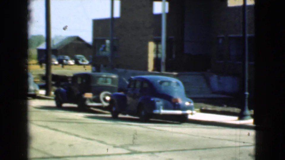 1939 Cleveland curbside view with a dark vintage car and blue sedan parked outside a brick building, seen through window bars.