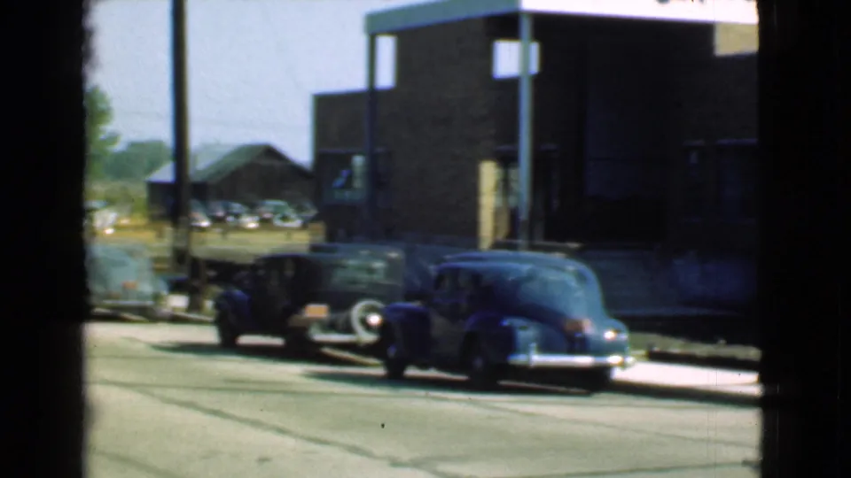 1939 Cleveland home-movie frame showing parked vintage cars beside a brick building, with more cars faintly visible farther back.