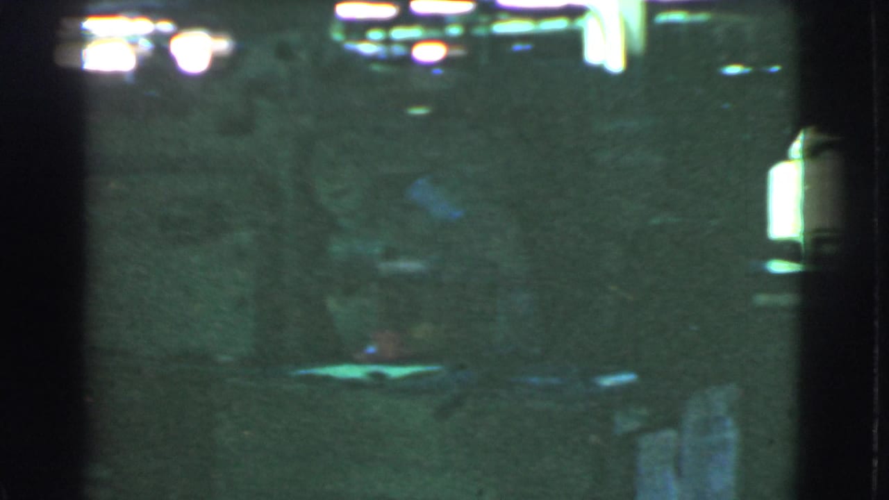 Dark green-tinted scene with blurred lights, shadows, and indistinct figures near machinery.