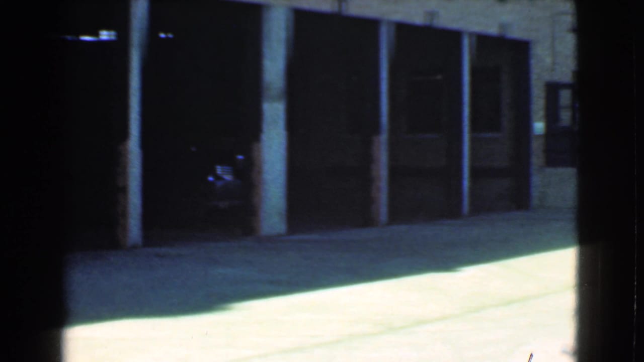 1939 Cleveland home-movie frame showing dark garage openings, brick walls, posts, and bright concrete pavement in an alleyway.