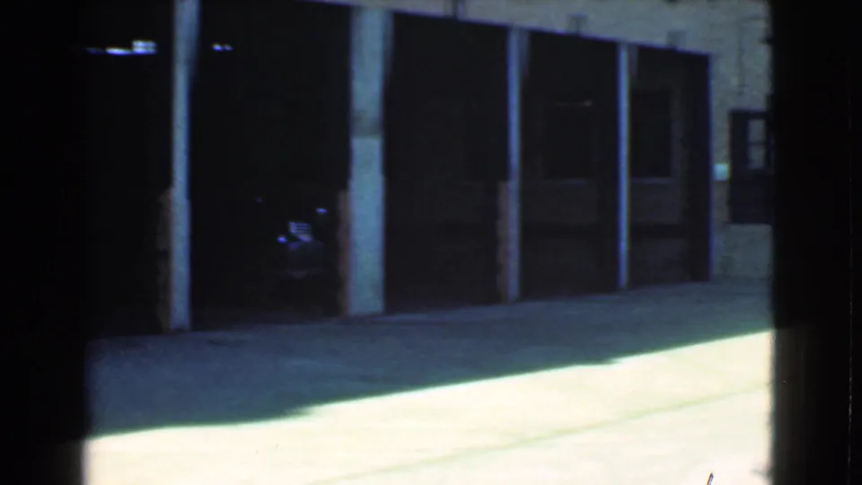 1939 Cleveland home-movie frame showing dark garage openings, brick walls, posts, and bright concrete pavement in an alleyway.