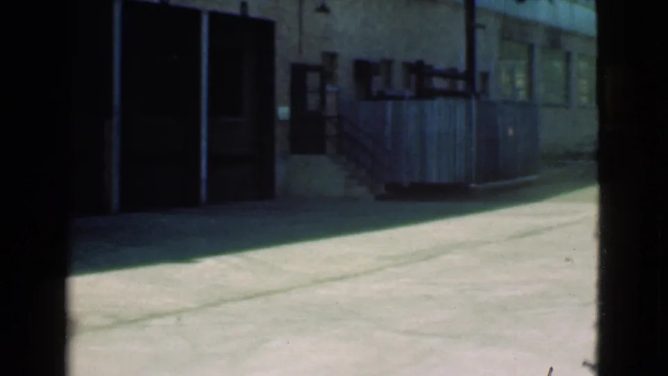 Cleveland alley frame showing a shadowed garage door, small windows, wooden fencing, exterior stairs, and sunlit concrete pavement.