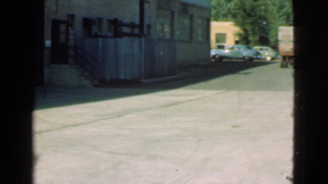 Street view with shaded building wall, parked vintage cars, trees, and a small lorry farther back.