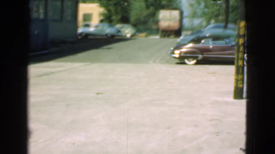 1939 Cleveland street view with a dark vintage car at right, a small lorry ahead, parked cars, trees, and a partial sign edge.