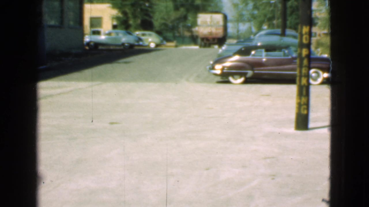 Street view with parked period cars, a truck, pavement, and a yellow no-parking pole.