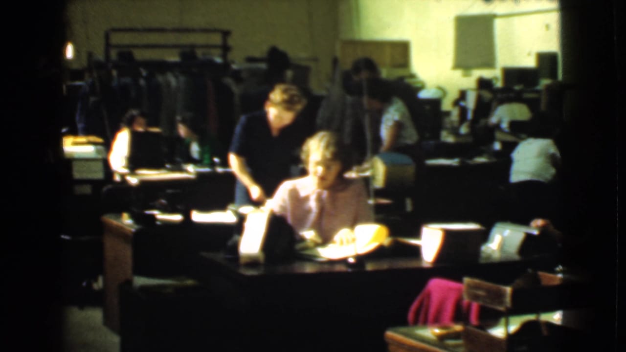 Women work at desks in a dim factory office with clothing racks behind them.