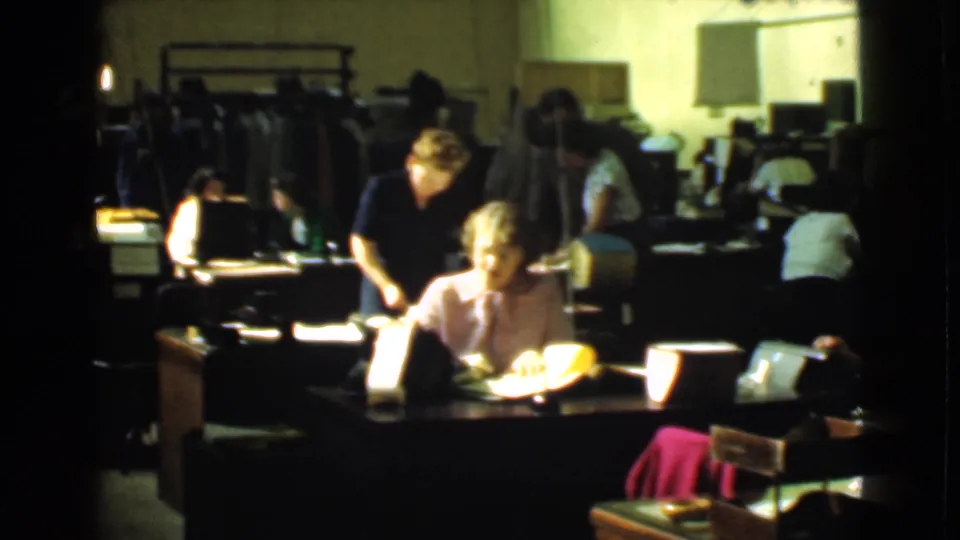 In Cleveland, Ohio, 1939, women work at desks in a dim factory office with papers, desk lamps and clothing racks in the background.