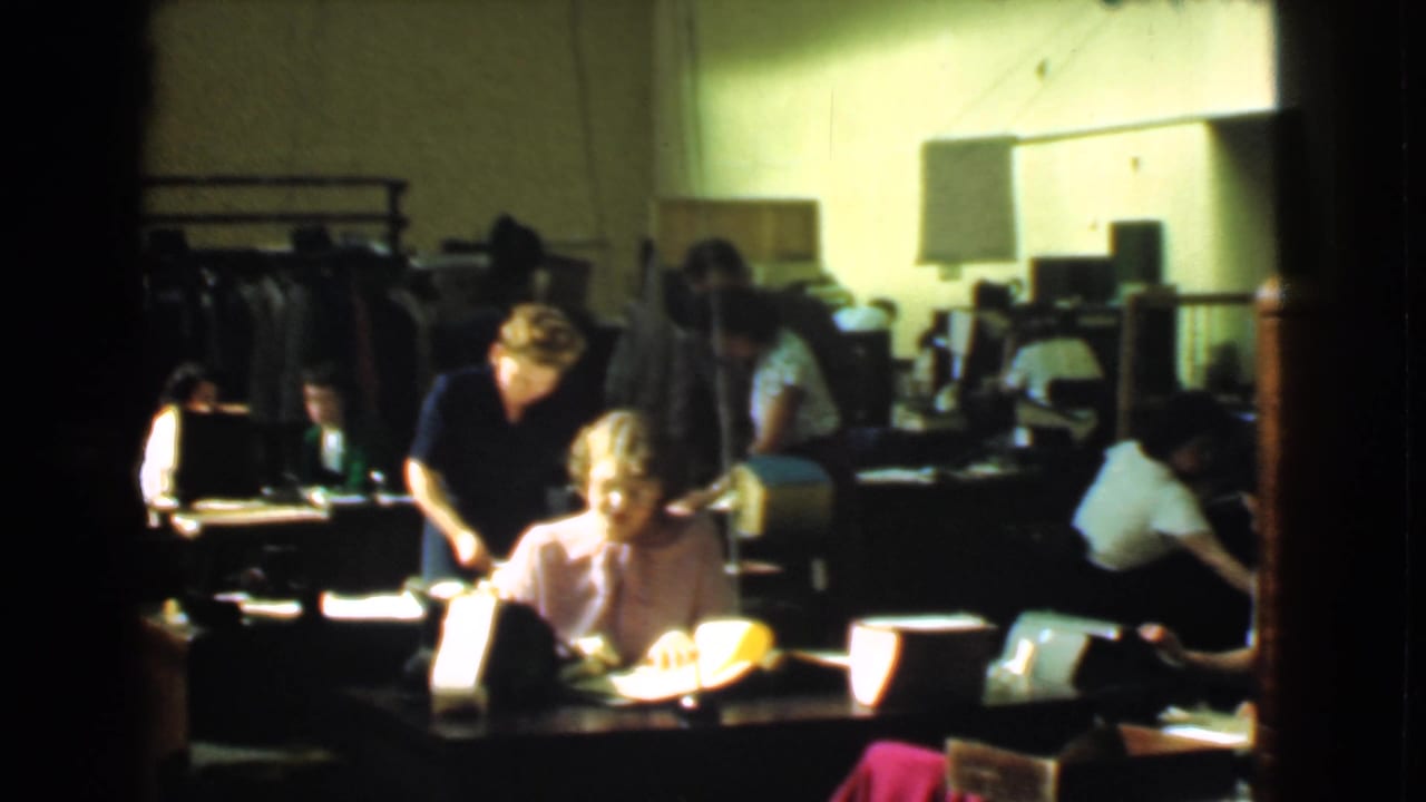 A seated woman handles papers at a desk while another woman stands nearby in the 1939 Cleveland factory office scene.