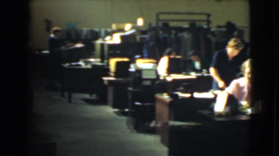 A wider view of the Cleveland factory office shows rows of desks, bright task lamps, clothing racks and women working at the right edge.