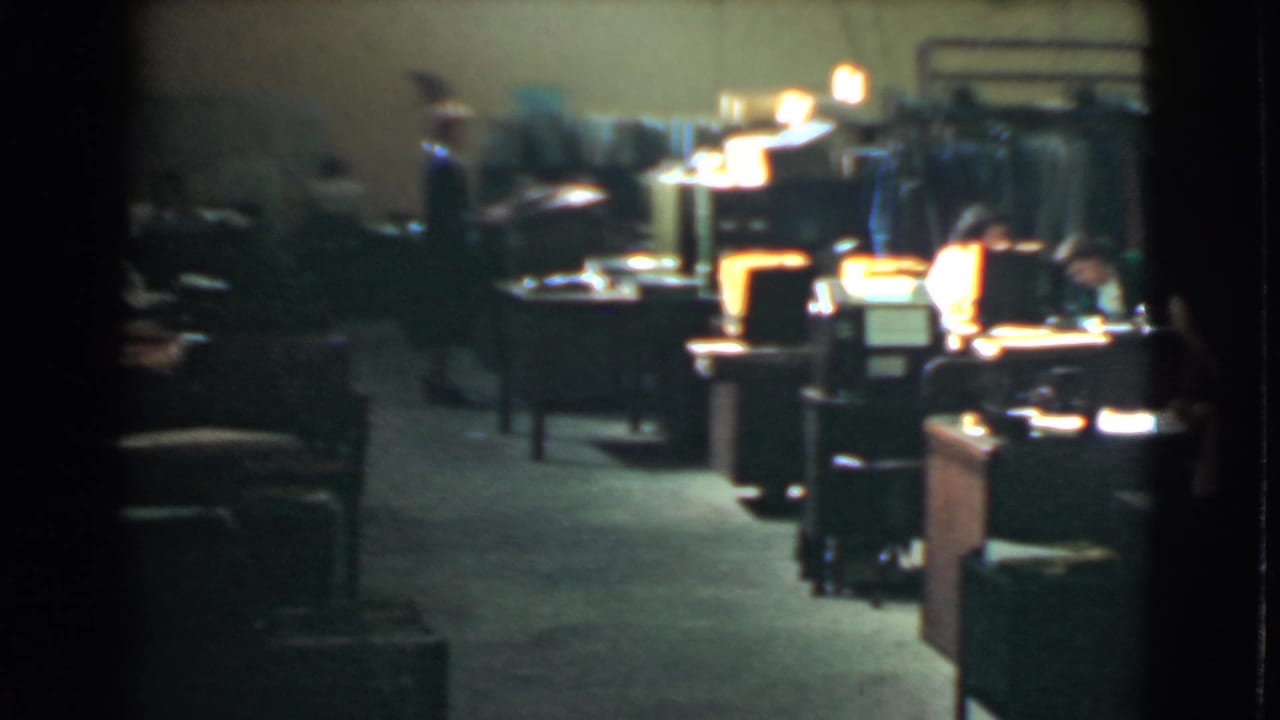 Dim office room with desks, chairs, lights, and workers in the background.