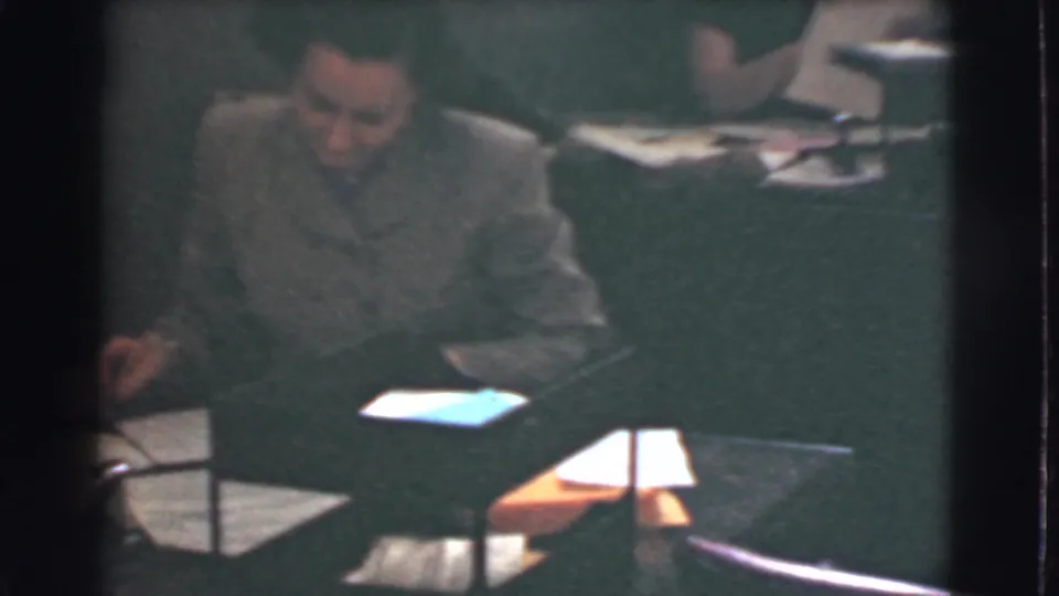 A man in period office attire sits beside desks, papers, and lamps in a 1939 Cleveland office interior.