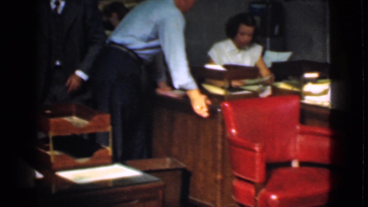 Man leans over a desk near a red chair while a woman works in the background.
