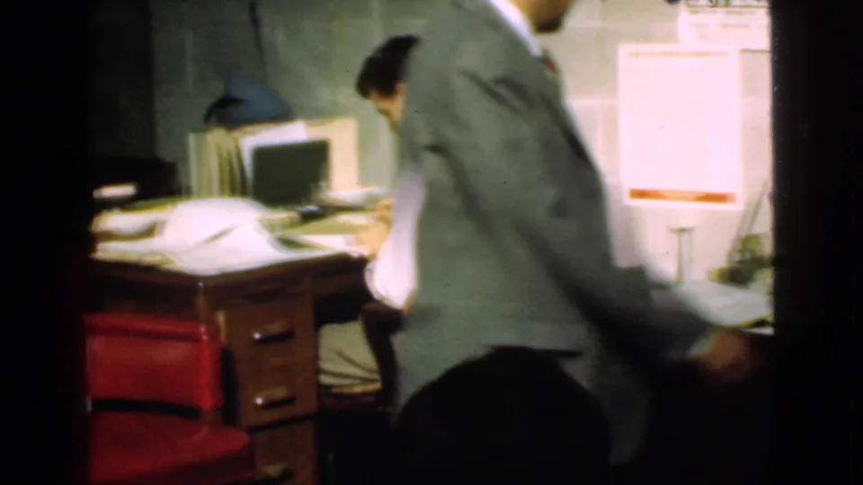 Cleveland office frame with a suited man crossing by a paper-covered desk, red chair, and small office machine.