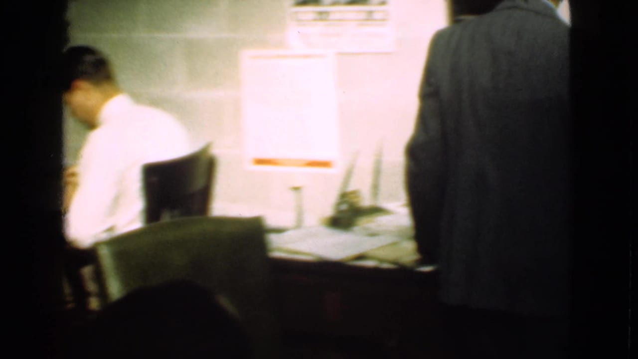 Man seated at a desk while another suited figure stands in a dim industrial interior.