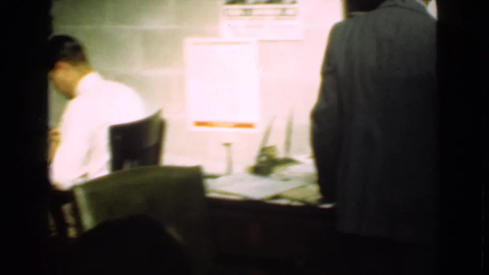 Cleveland factory interior from 1939 with a seated man at a desk, wall notices, and another figure partly blocking the corridor view.