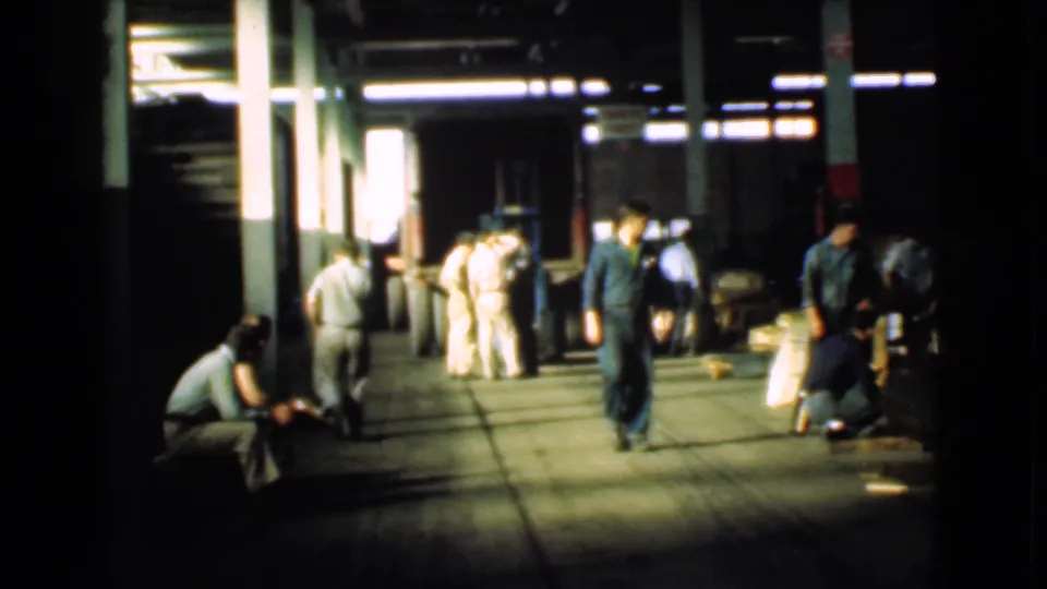 A man in a green jumpsuit walks through a 1939 Cleveland factory corridor while workers in light uniforms gather farther back.