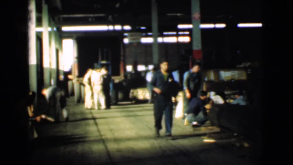 The green-clad worker continues through the Cleveland factory corridor, with men crouching near the floor and others gathered by columns.